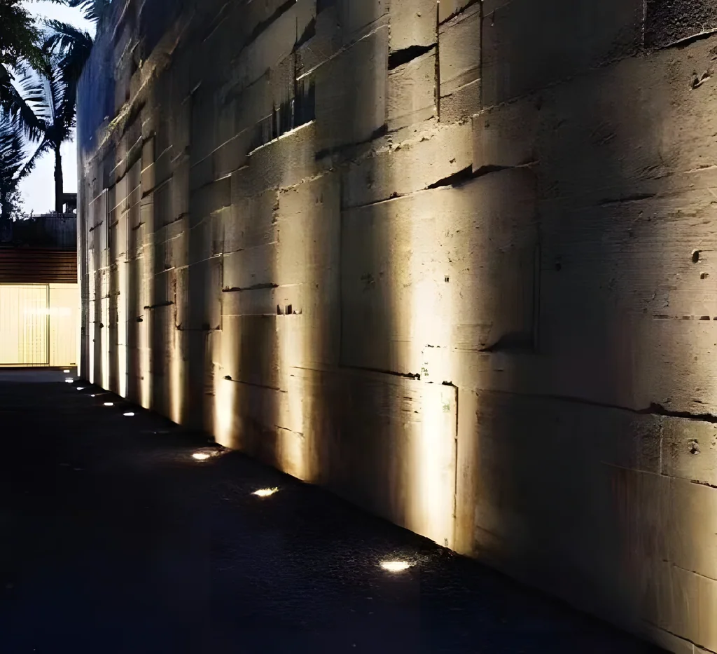 Illustrative application photo: Round in-ground uplights grazing a textured stone wall at night.