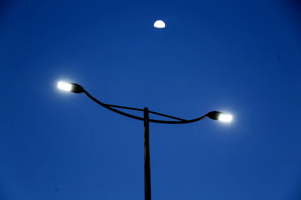 Two luminaire heads on a double-arm pole, lit against a deep blue evening sky with the moon above