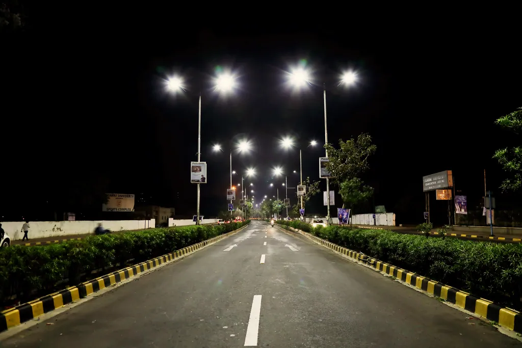 An empty road at night lit by double-arm street lights, with hedged medians and painted kerbs