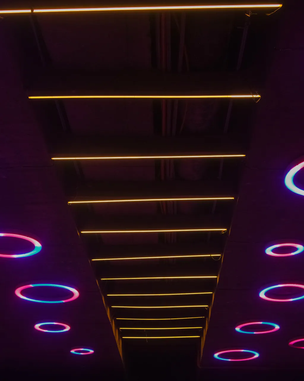 Looking up at the central beam of the bridge, crossed by warm lines of light and flanked by coloured rings