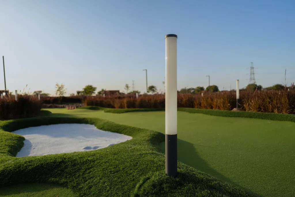White tube bollard with a dark base standing on a green beside a sand bunker