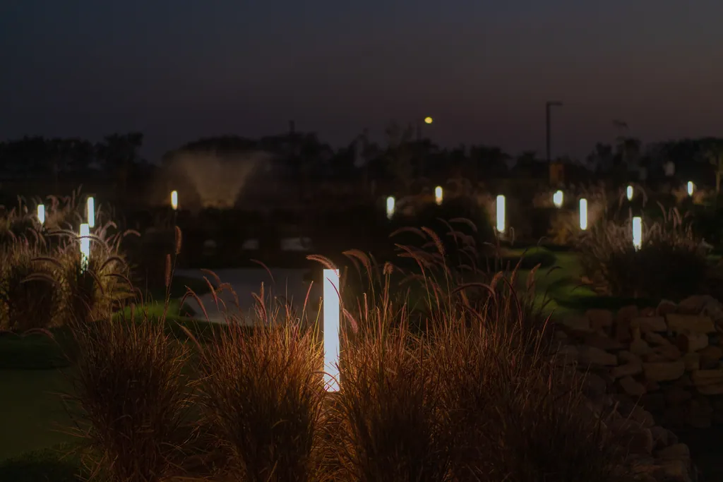 Tube bollard glowing through grasses after dark, with a fountain spraying in the background