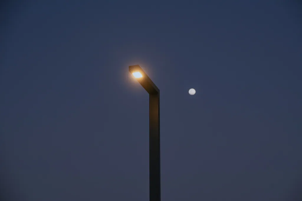 Single-arm pole lit at dusk, with the moon in the sky beside it