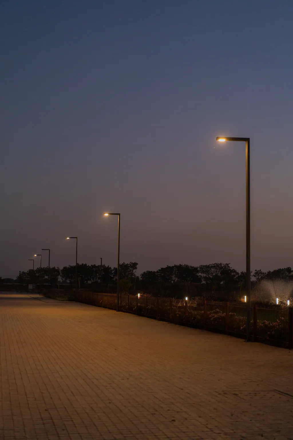 Promenade at dusk lit by a line of single-arm poles, with bollards along the fence